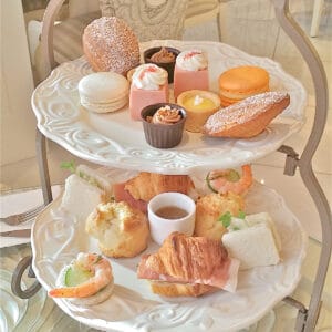 A 2-tier afternoon tea display featuring little cute treats in pastel colors and little croissant sandwiches.