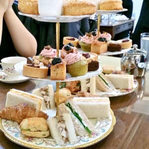Two afternoon tea sets filled with mini desserts and sandwiches on a wooden tabletop.