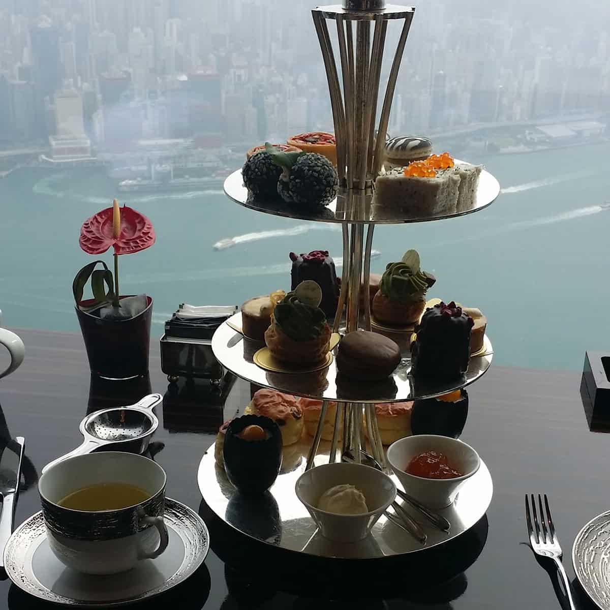An afternoon tea set on a 3-tier silver tray in front of a window overlooking Hong Kong harbor.