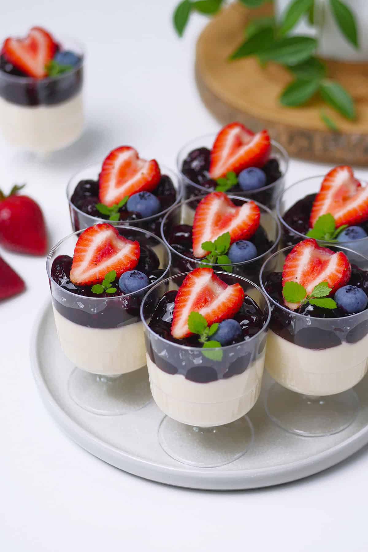 A group of mini cups of panna cottas are laid out on a round serving plate. The mini desserts are topped with mixed berry sauce and fresh berries like strawberries and blueberries.  
