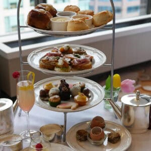 A traditional 3-tier afternoon tea set laid out in front a of window with views of tall buildings in downtown Vancouver.