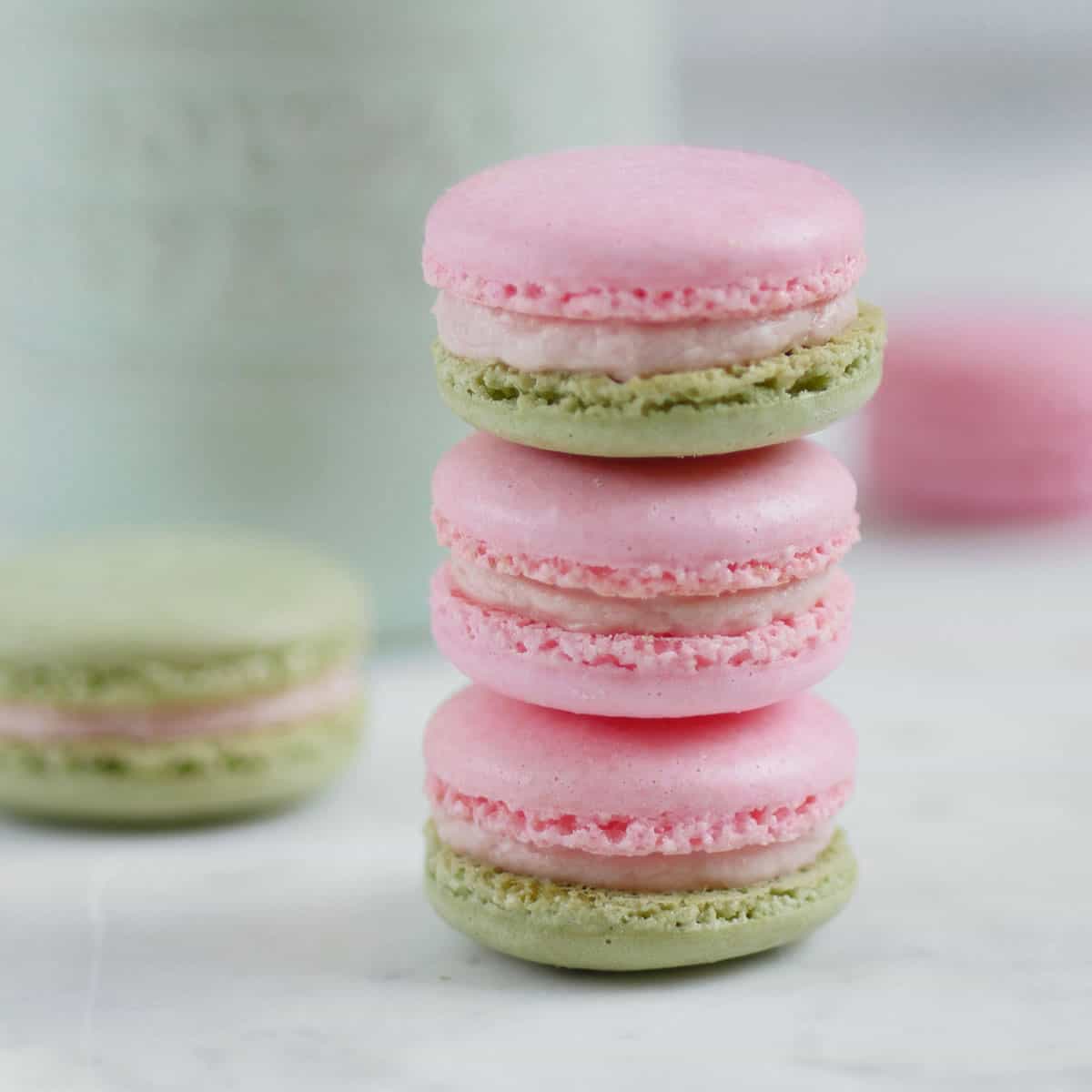 A stack of strawberry basil macarons with green and pink shells in front of a cup of strawberries.
