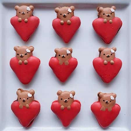 Macarons in the shape of bears hanging onto red hearts. 