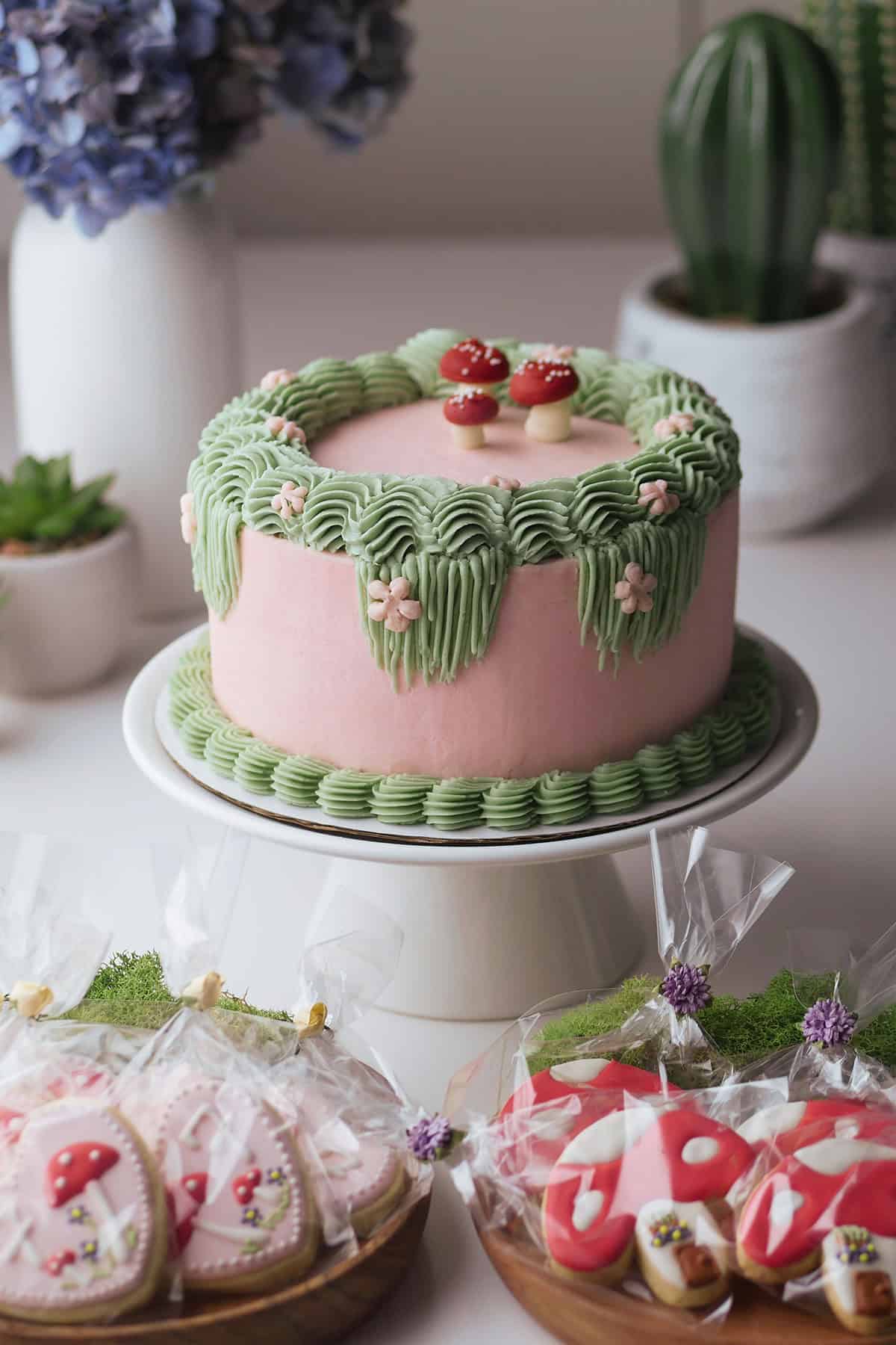 A full profile of a fairy themed cake on a cake stand. Below it are forest themed sugar cookies on a bed of moss. 