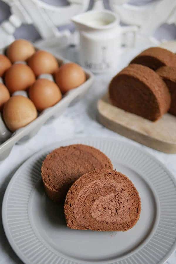 Chocolate cake roll slices on a grey plate with eggs and a creamer in the back. 