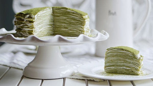 Mille crepe cake on a cake stand with a piece on the side. Both shows 28 layers of crepes stacked to make mille crepe cake.