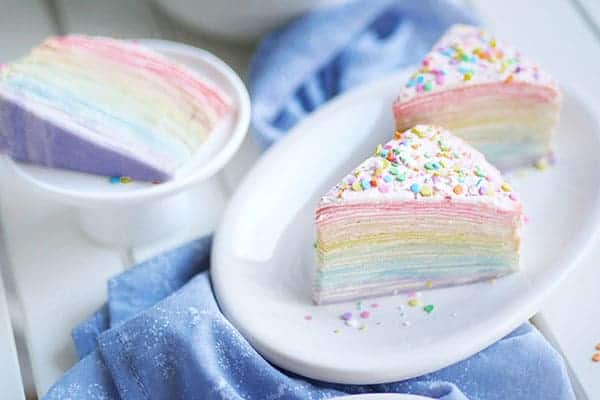 A top down look at 3 slices of rainbow colored crepe cakes with sprinkles on top laid out on 2 dishes. 