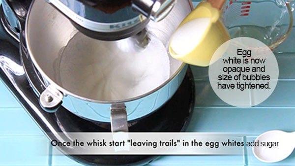 Opaque egg whites in a counter top mixer while sugar is being poured in. 