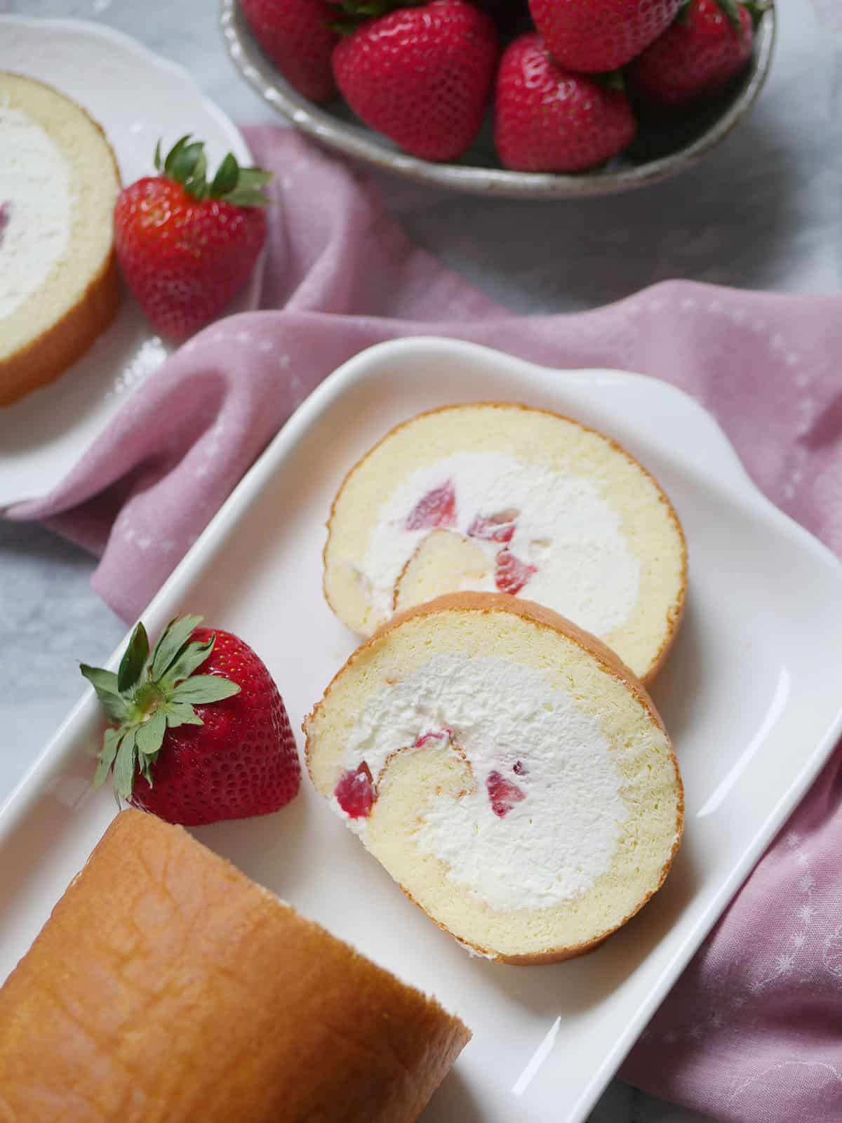 Strawberry cake roll with a few slices cut out and strawberries on a bowl in the back. 