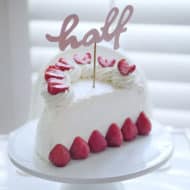 A half birthday cake made from whipped cream and fresh strawberries topped with a cake topper that says "half".