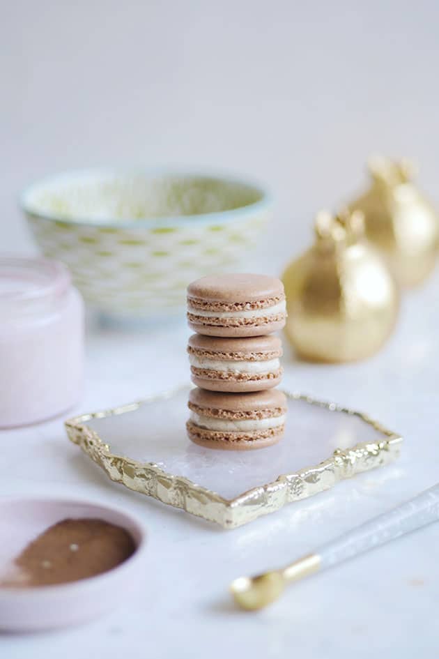 A stack of gingerbread spice macarons on an agate place set in front of a bowl, gold shakers and a candle and behind a plate of spice. 