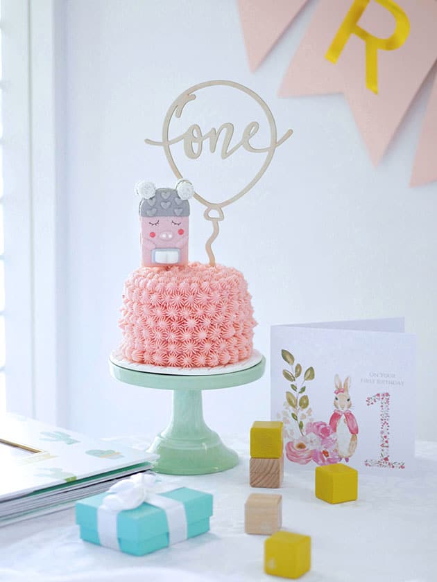 A mini pink smash cake on a green cake stand placed on a table with kid's wooden blocks and a birthday card. 