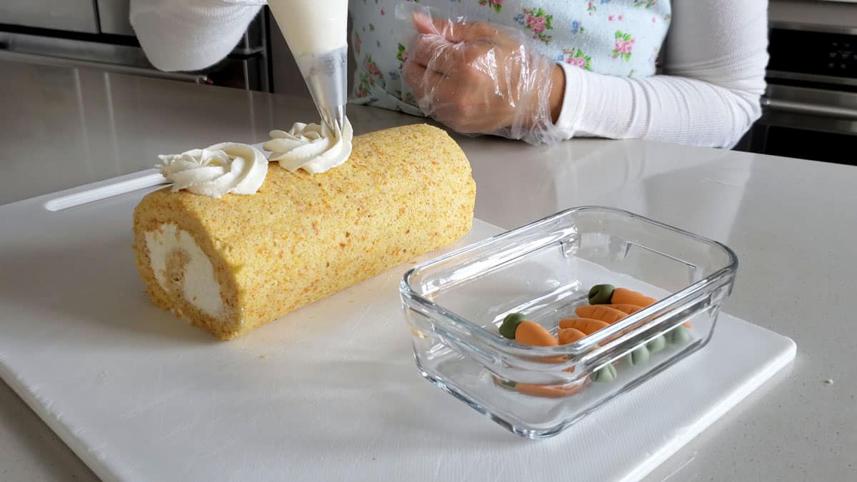 Marscapone cream being piped onto the top of the roll cake sitting on a white cutting board.