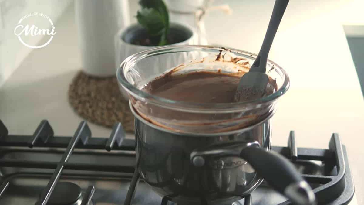 A bowl of melted chocolate cream is hanging over a pot of hot water on a stove top. 