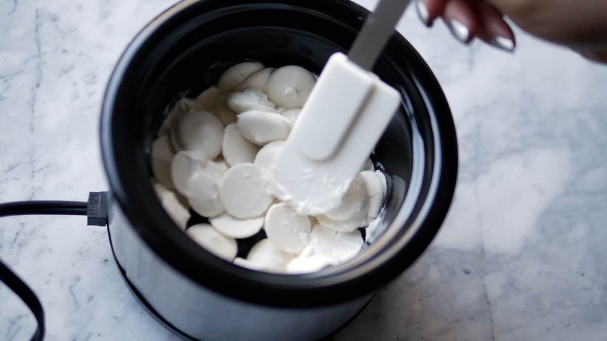 A hand stirring white candy melts in a mini crockpot with a small spatula. 