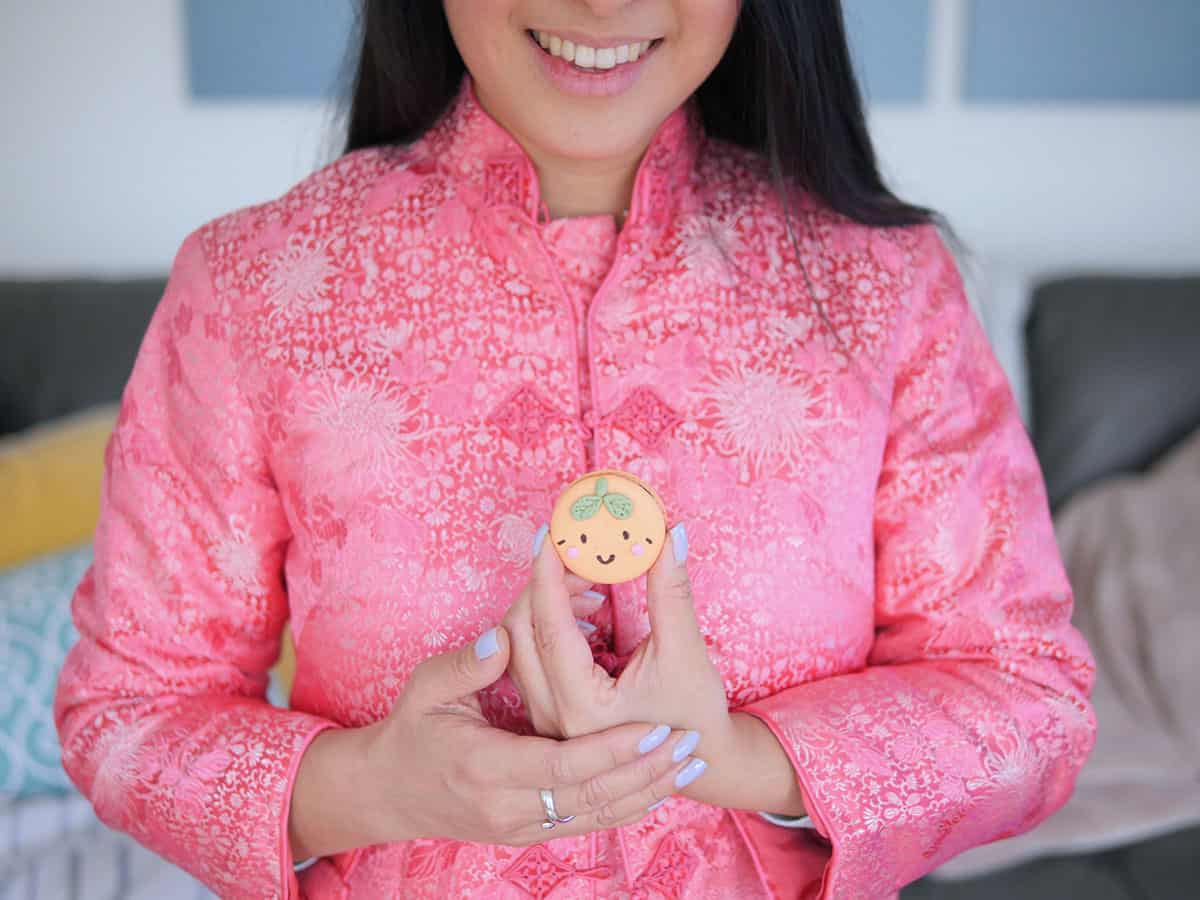 Hand holding a mandarin orange macaron with Chinese new years attire in the background.