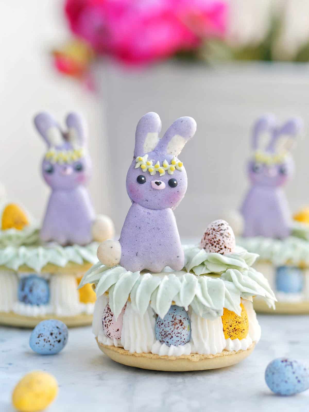Three sets of Easter bunnies perched on top of a big macaron filled with Easter eggs. 