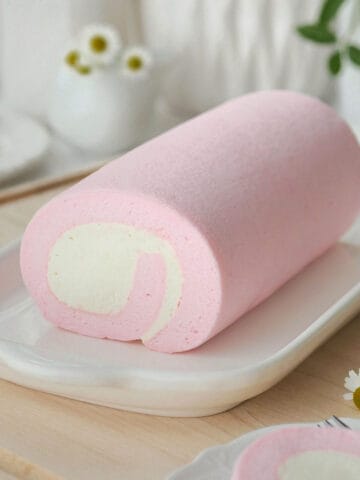 Pink cotton candy cake roll on a long plate on table adorned with white flowers.