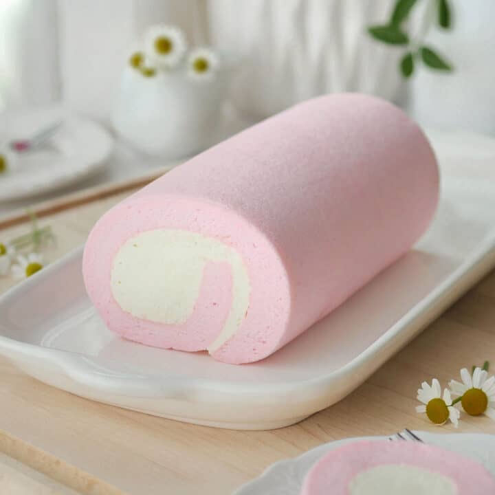 Pink cotton candy cake roll on a long plate on table adorned with white flowers.