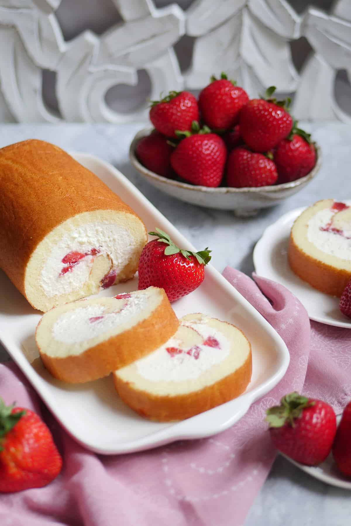 Table top with fresh strawberries in the back and swiss roll in the foreground. 