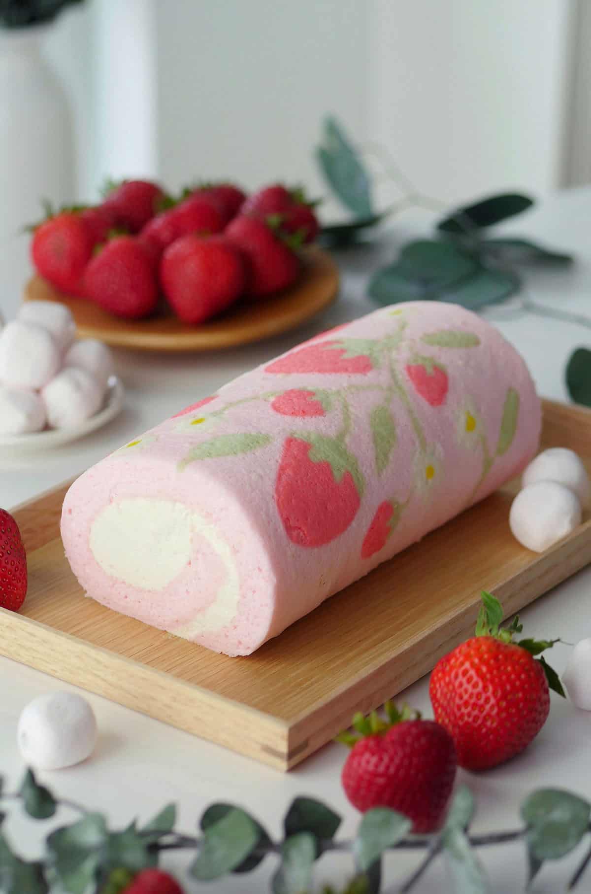 Cake roll with intricate pattern on a wooden tray with strawberries in the back. 