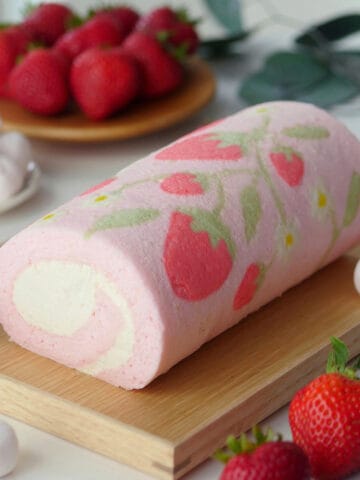 Pink cake roll decorated with strawberry print resting on a wooden tray with fruits in the back.