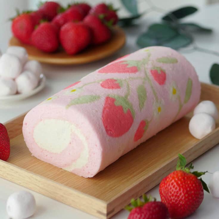 Pink cake roll decorated with strawberry print resting on a wooden tray with fruits in the back.