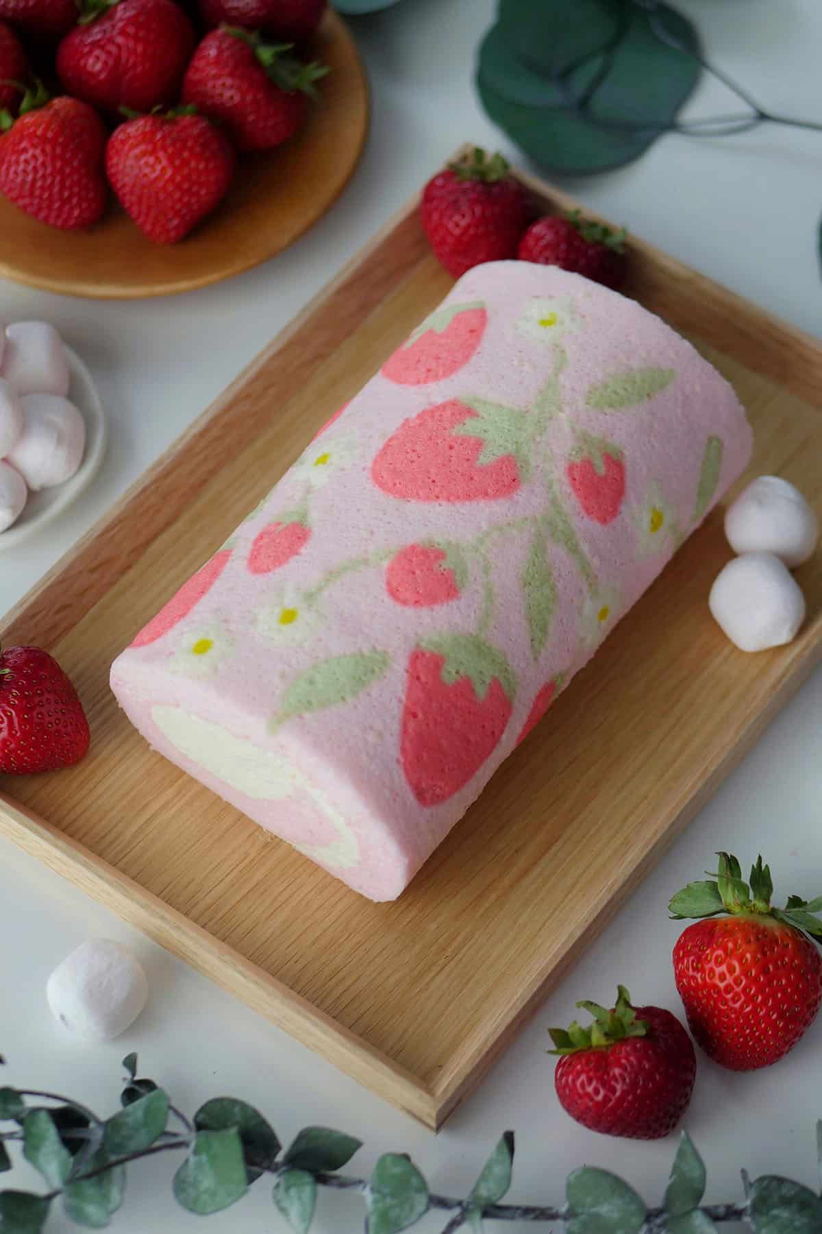 A pink cake roll on a wooden plate with strawberries and marshmallows on the side. 