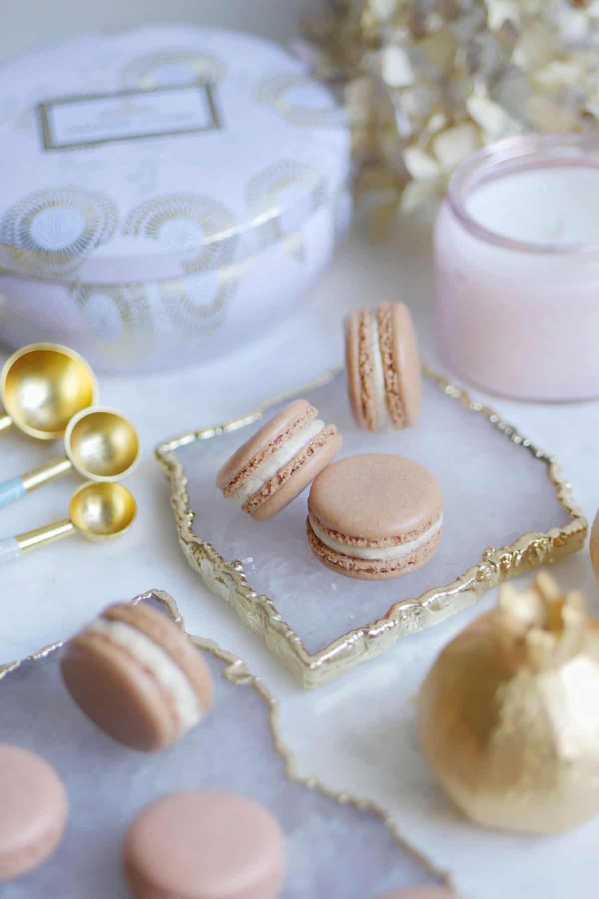 A bunch of gingerbread spice macarons amongst a variety of kitchen utensils and plates with gold trim. 