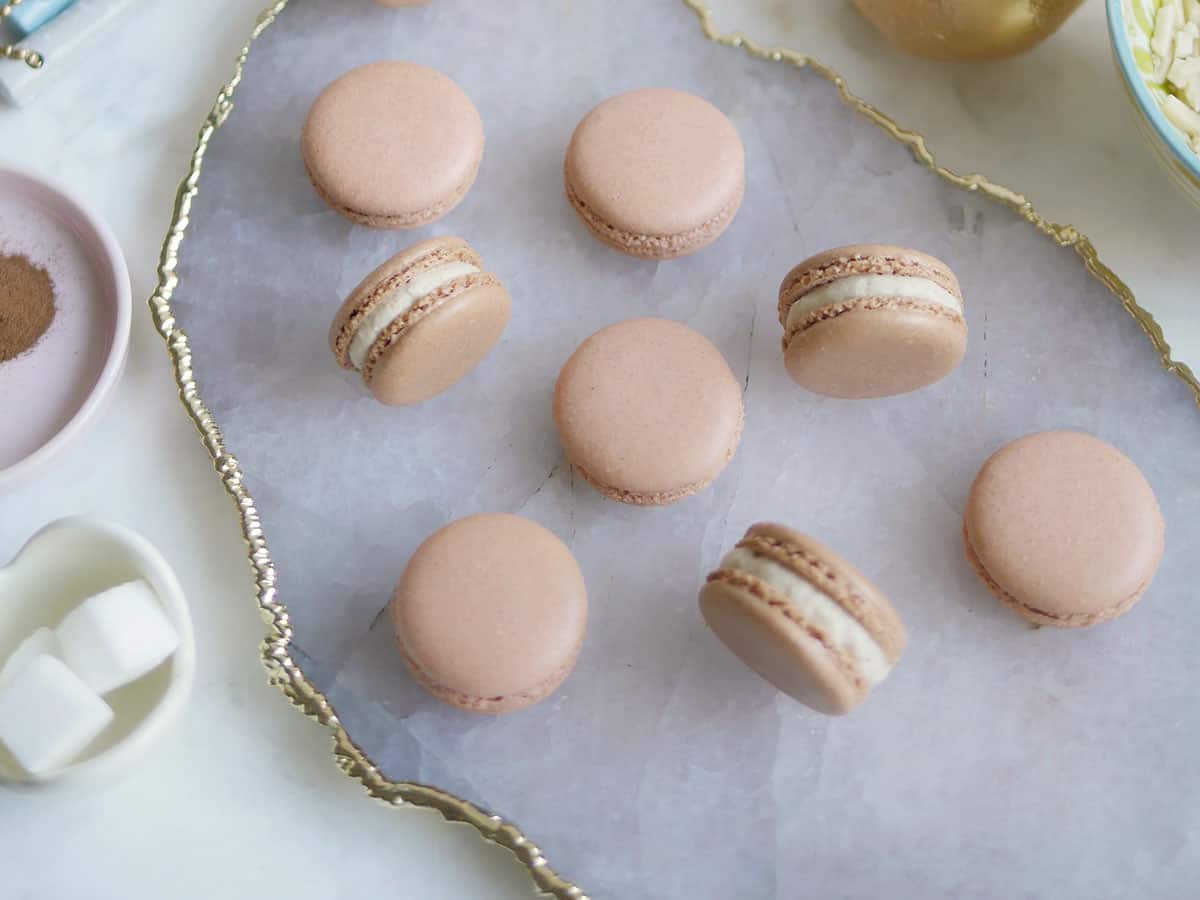 Bird's eye view of many gingerbread macarons on an agate plate trimmed with gold. Plates of of spice and sugar flank the sides. 