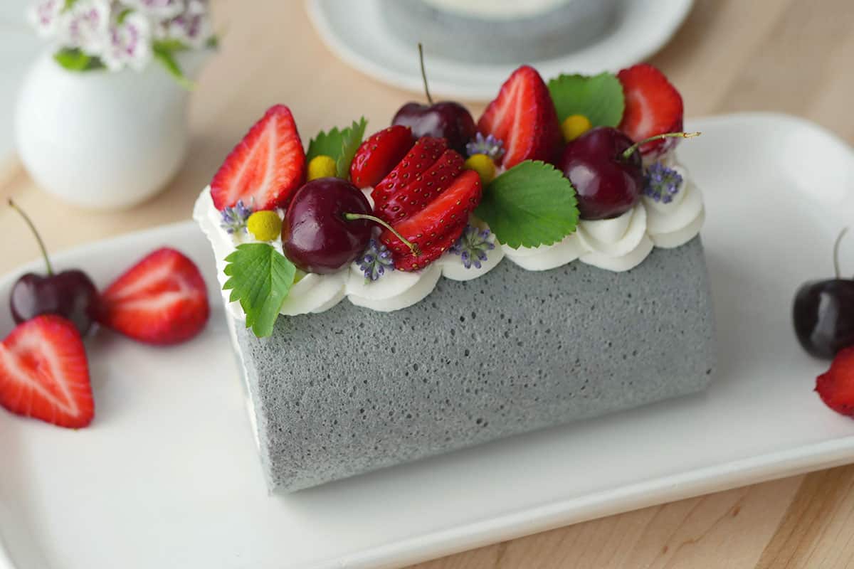 A beautiful grey colored Earl Grey cake roll on a white plate topped with fresh cream and a variety of flowers and fruits. 