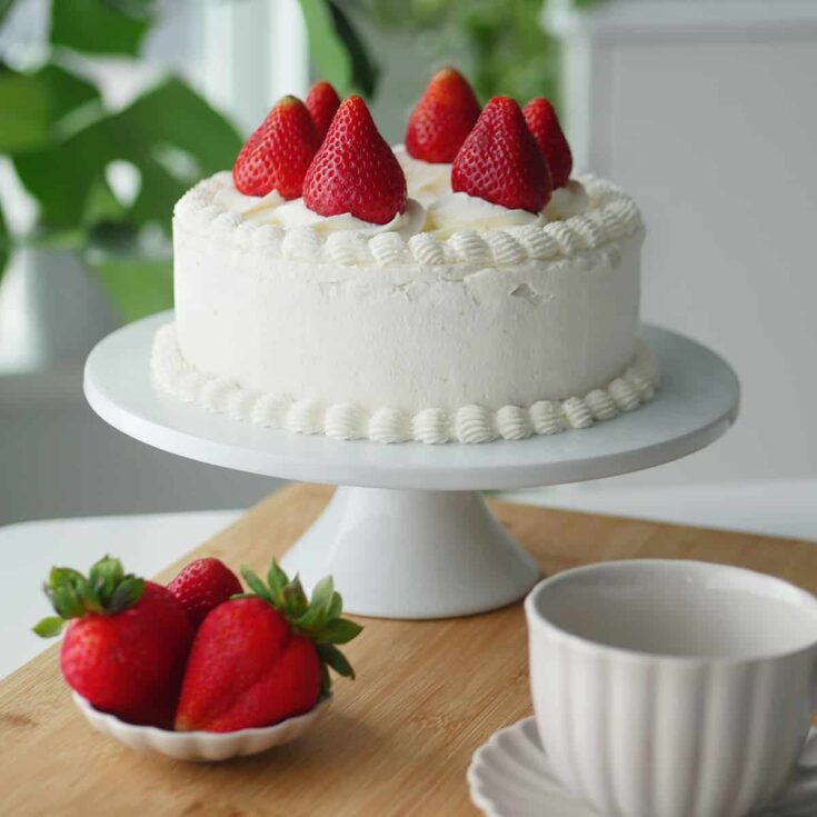 Fresh fruit cream cake on a white cake stand in a sunlit dining room.