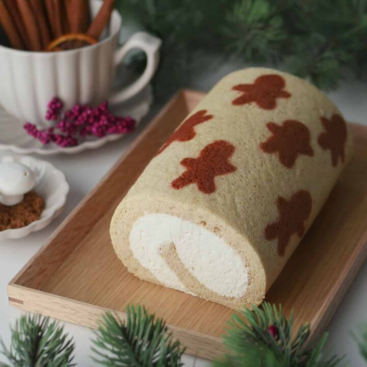 Gingerbread men pattern cake roll on a wooden serving plate with cinnamon and spices in the back.