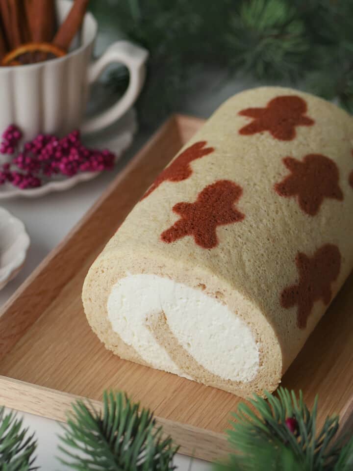 Gingerbread men pattern cake roll on a wooden serving plate with cinnamon and spices in the back.