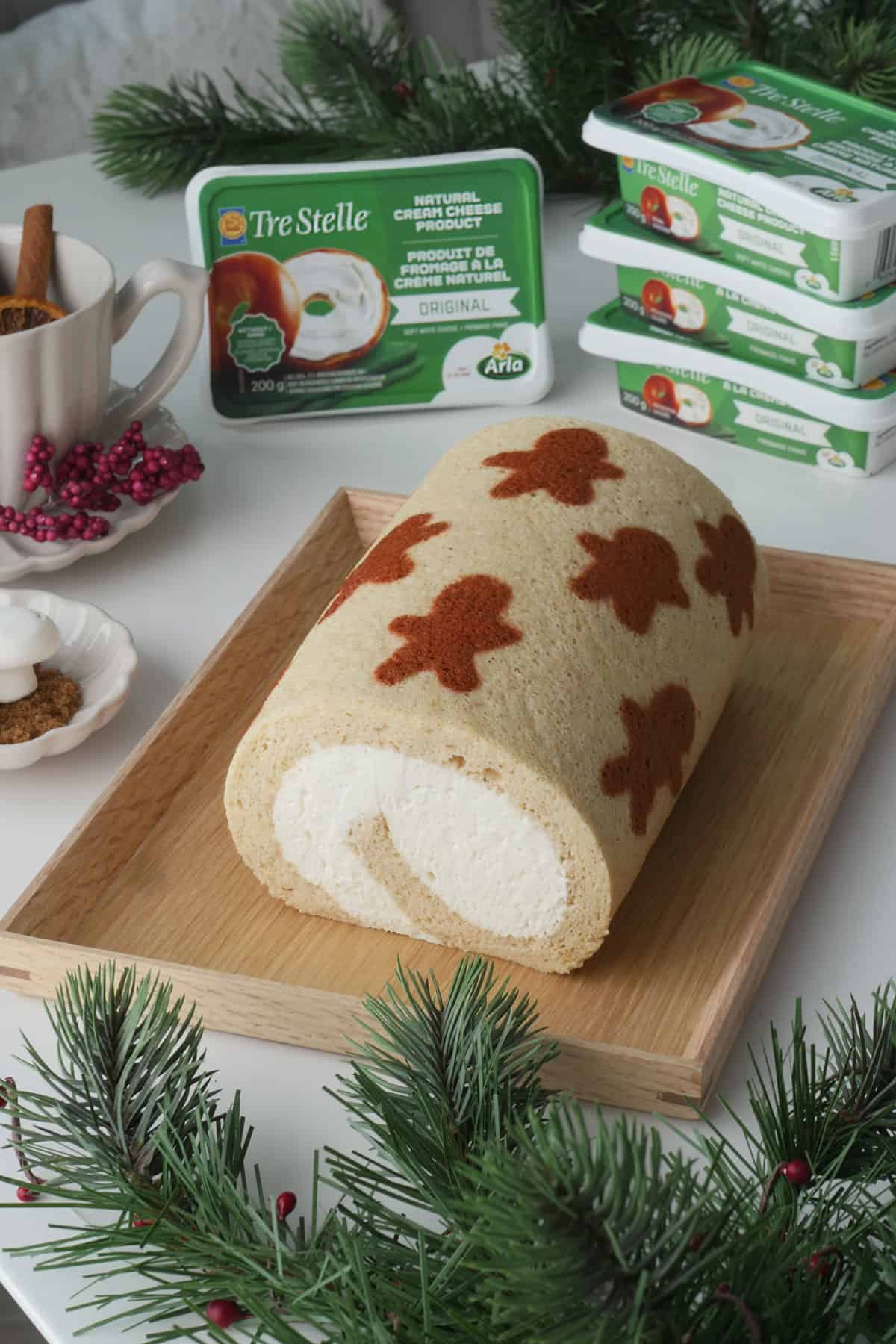 A festive cake roll with holiday patterns printed on it is on a wooden serving tray with some cream cheese packets in the background.