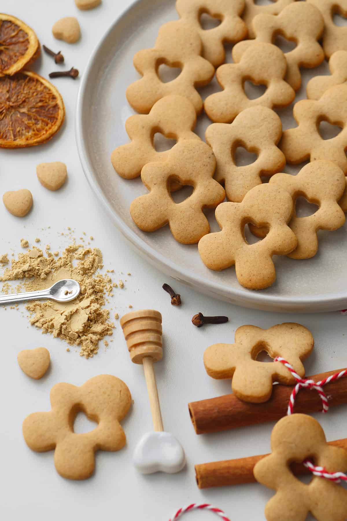Gingerbread men are on a round plate with festive dried orange slices, honey stick, cinnamon sticks surrounding it. 