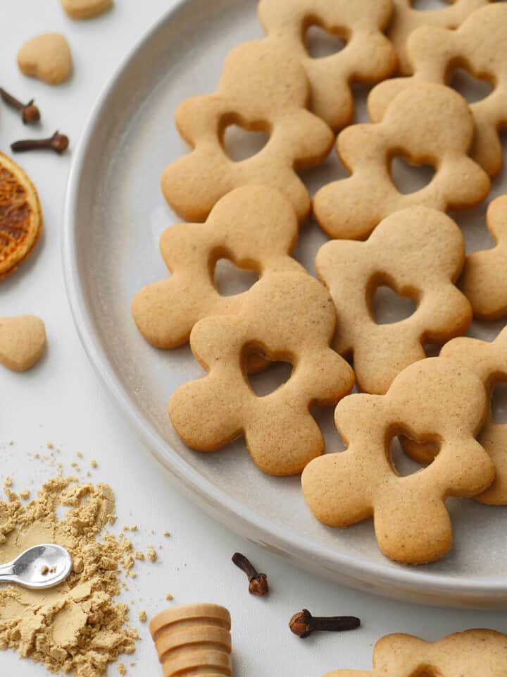 Honey gingerbread spice cookies cut in gingerbread shape with a heart cutout. All laid out on a stoneware plate with golden spices on the side.