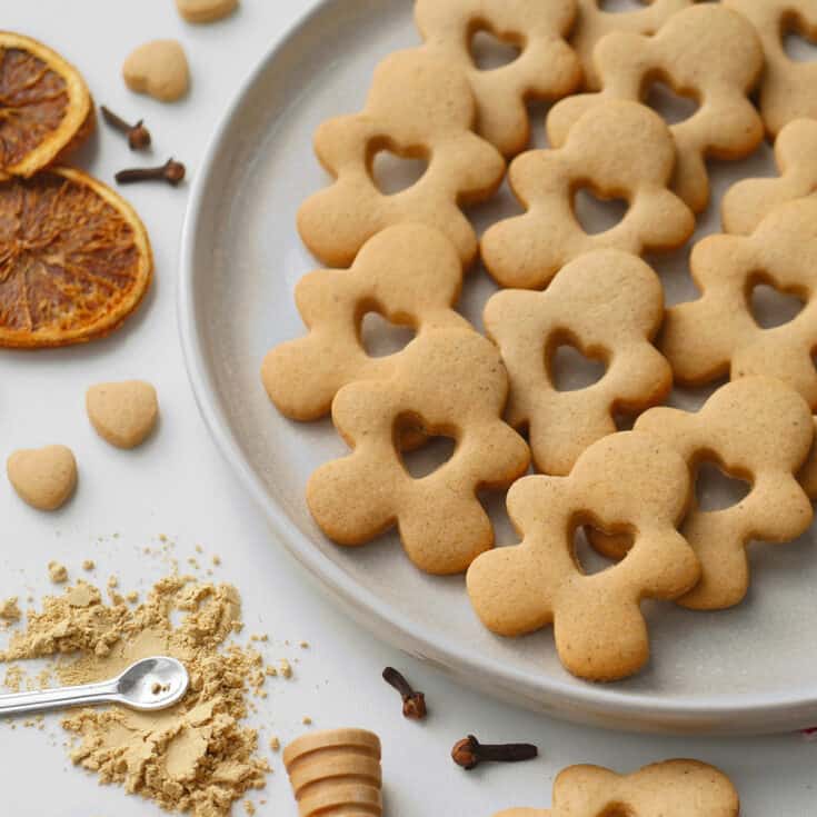 Honey gingerbread spice cookies cut in gingerbread shape with a heart cutout. All laid out on a stoneware plate with golden spices on the side.