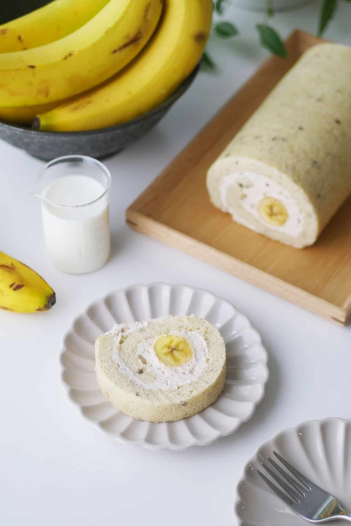 On a white table rests a banana cake roll on a plate with a bowl of fresh bananas and a glass of milk next to it. 