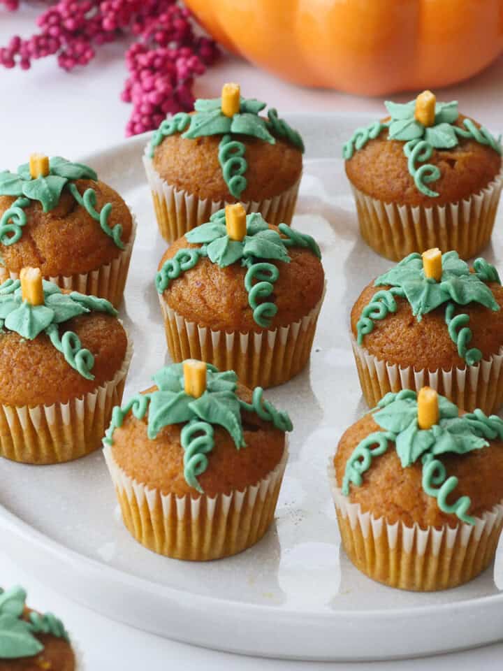 Many mini pumpkin cupcakes decorated with buttercream to look like pumpkin leaves on a round plate.