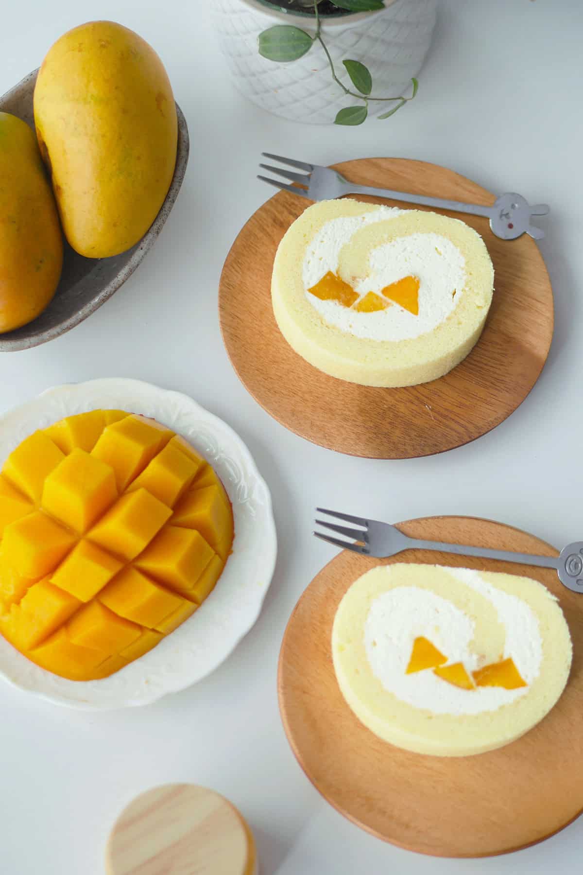 Two slices of mango cake roll on a wooden plate flanked by another plate with a slice of fresh mango that is partially cubed. 