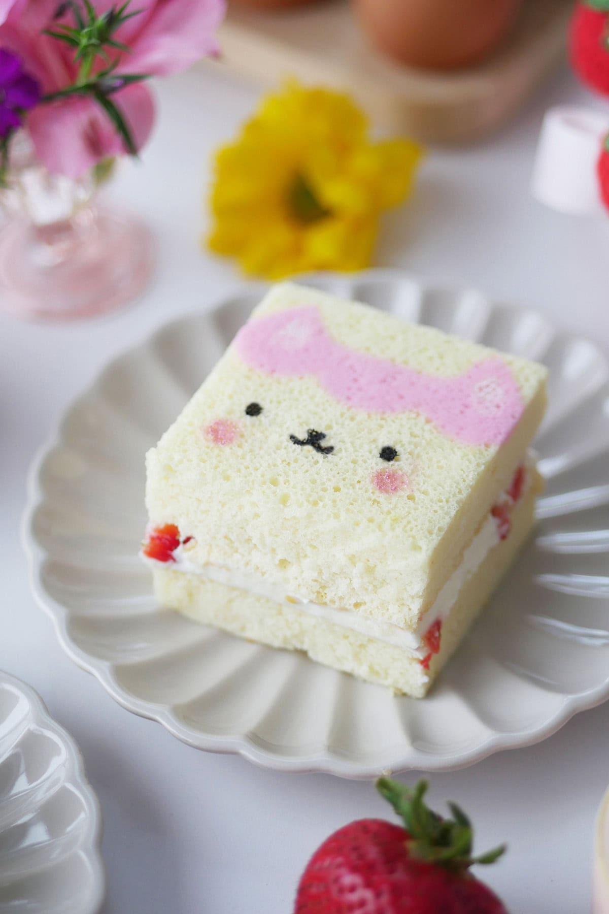 A slice of printed cake of a Gabby Dollhouse character sits on a scalloped plate surrounded by flowers.