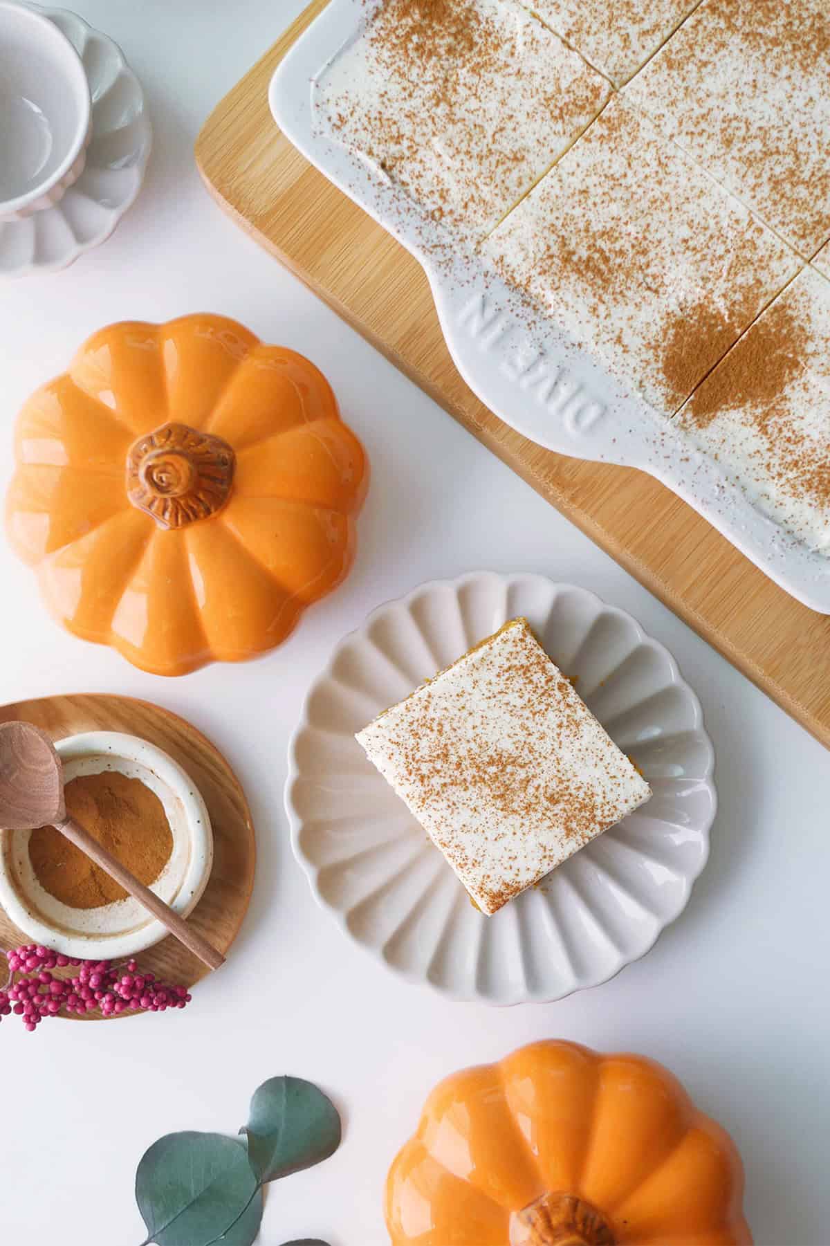 Top down view of Tres Leches cake slice on a plate surrounded by mini pumpkins and a plate of pumpkin spice with a wooden spoon resting on top. 