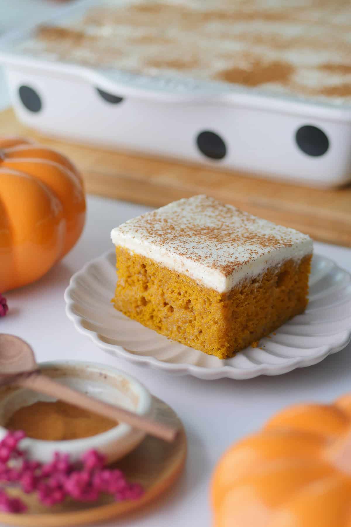 A close up of a square slice of pumpkin Tres Leches cake on a plate surrounded by little pumpkins and a pot of ground spices. 