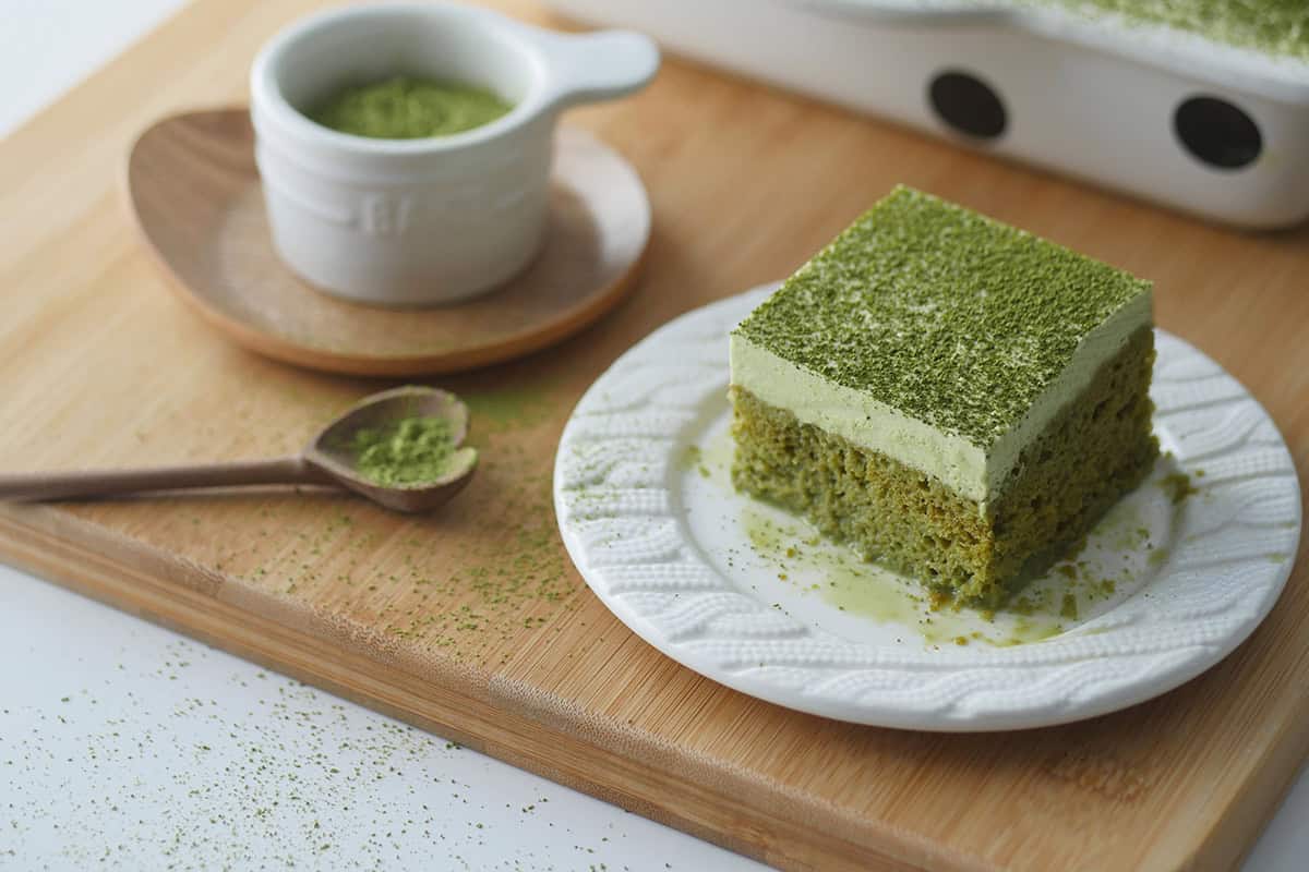 A slice of Matcha Tres Leches is shown on a small dessert plate, on the side of it is a small heart shaped wooden spoon filled with Matcha powder.