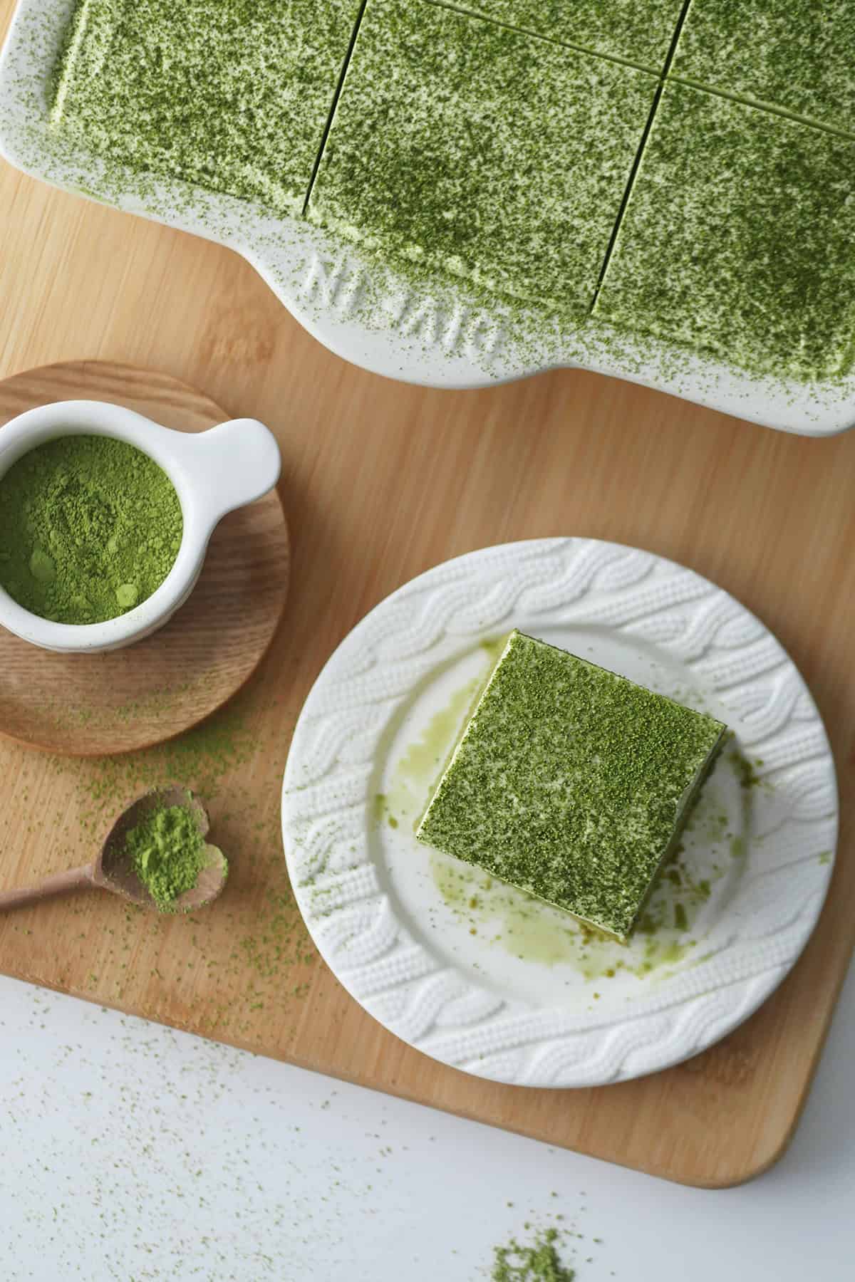 In the frame, half a pan of Matcha Tres Leches cake is shown along with a cup filled with green tea powder and a plate with one square slice of Matcha cake.