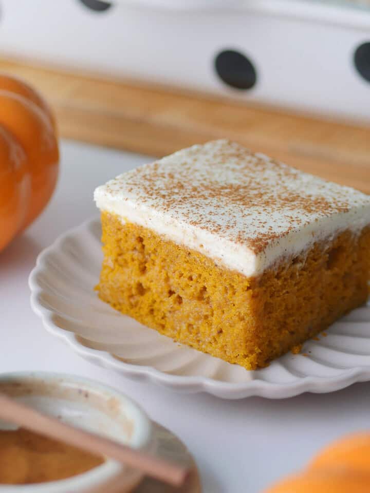 A square slice of pumpkin Tres Leches cake rests on a plate with a small pumpkin and a small bowl of spice next to it.