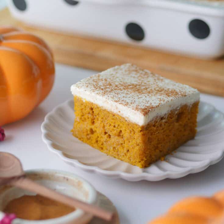 A square slice of pumpkin Tres Leches cake rests on a plate with a small pumpkin and a small bowl of spice next to it.