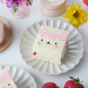 A slice of printed cake of a Gabby Dollhouse character sits on a scalloped plate surrounded by flowers.