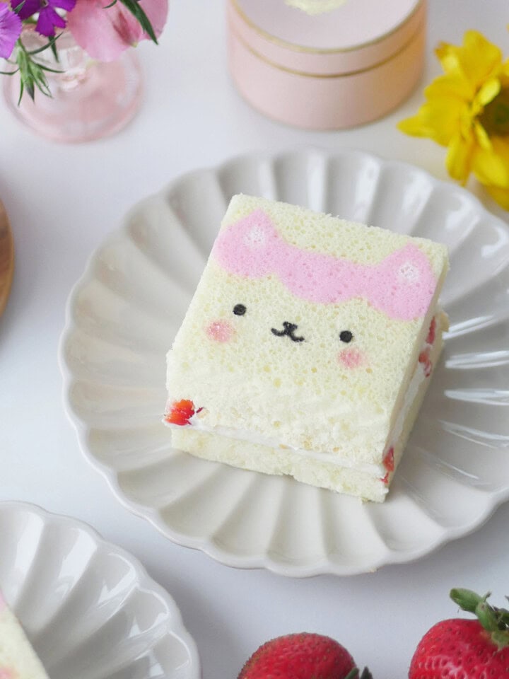 A slice of printed cake of a Gabby Dollhouse character sits on a scalloped plate surrounded by flowers.